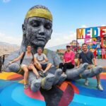 Turistas disfrutan de un tour privado en la Comuna 13 de Medellín, sentados en la icónica escultura gigante con vistas panorámicas y el letrero 'Medellín Es Una Chimba' al fondo. Representa la transformación social y el arte urbano.