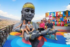 Turistas disfrutan de un tour privado en la Comuna 13 de Medellín, sentados en la icónica escultura gigante con vistas panorámicas y el letrero 'Medellín Es Una Chimba' al fondo. Representa la transformación social y el arte urbano.