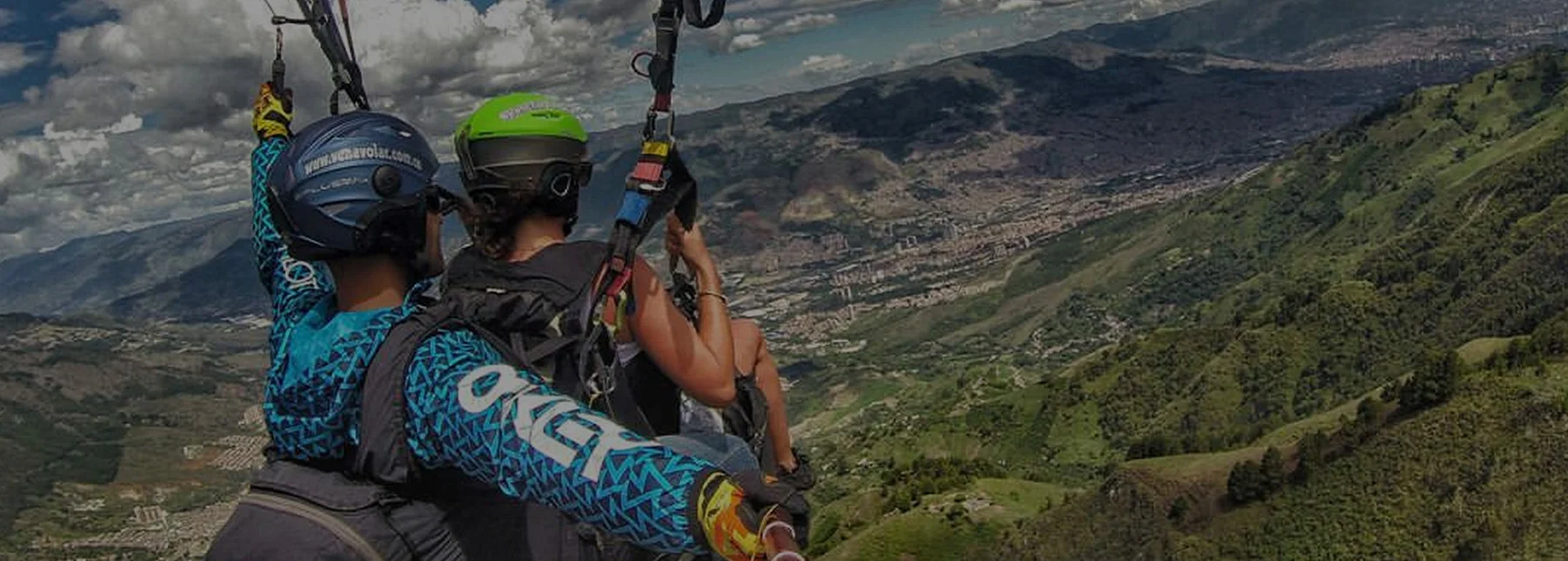 Vista en primer plano de turistas haciendo parapente tándem con piloto experto, sobrevolando las montañas verdes de San Félix y la ciudad de Medellín al fondo. Representa un tour de aventura aéreo seguro.