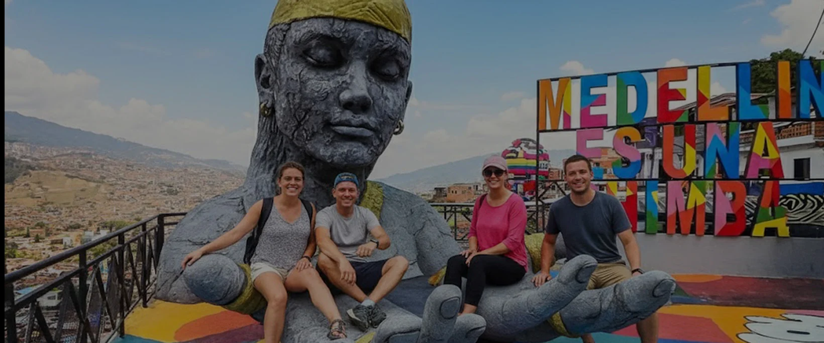 Turistas disfrutando de un tour privado en la Comuna 13 de Medellín, sentados en la icónica escultura de transformación social con vistas panorámicas de la ciudad y el cartel 'Medellín Es Una Chimba'